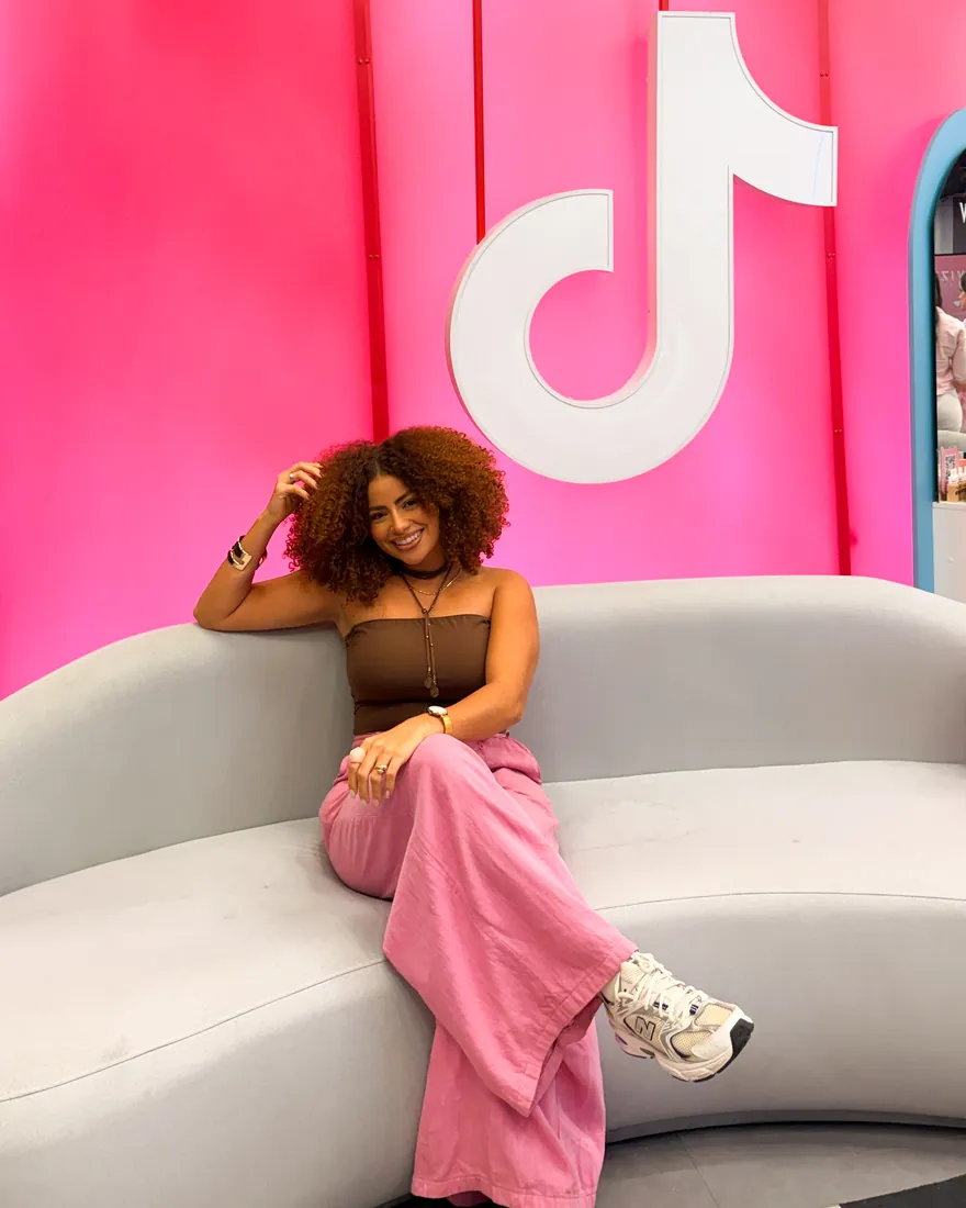 Esther Custódio sentada num sofá, sorrindo, num estúdio rosa com o logo do TikTok